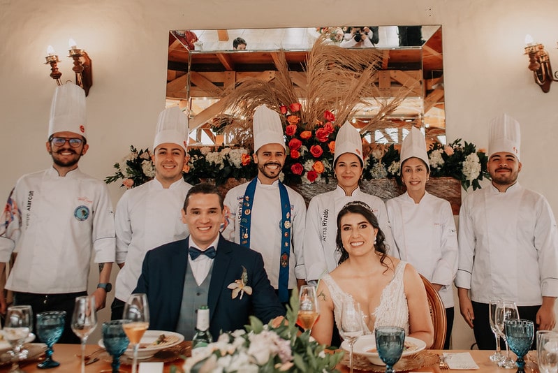 Pareja de recién casados sentados en la mesa nupcial, posando felices con el equipo de chefs que preparó el catering, en una boda decorada con flores y estilo rústico elegante.