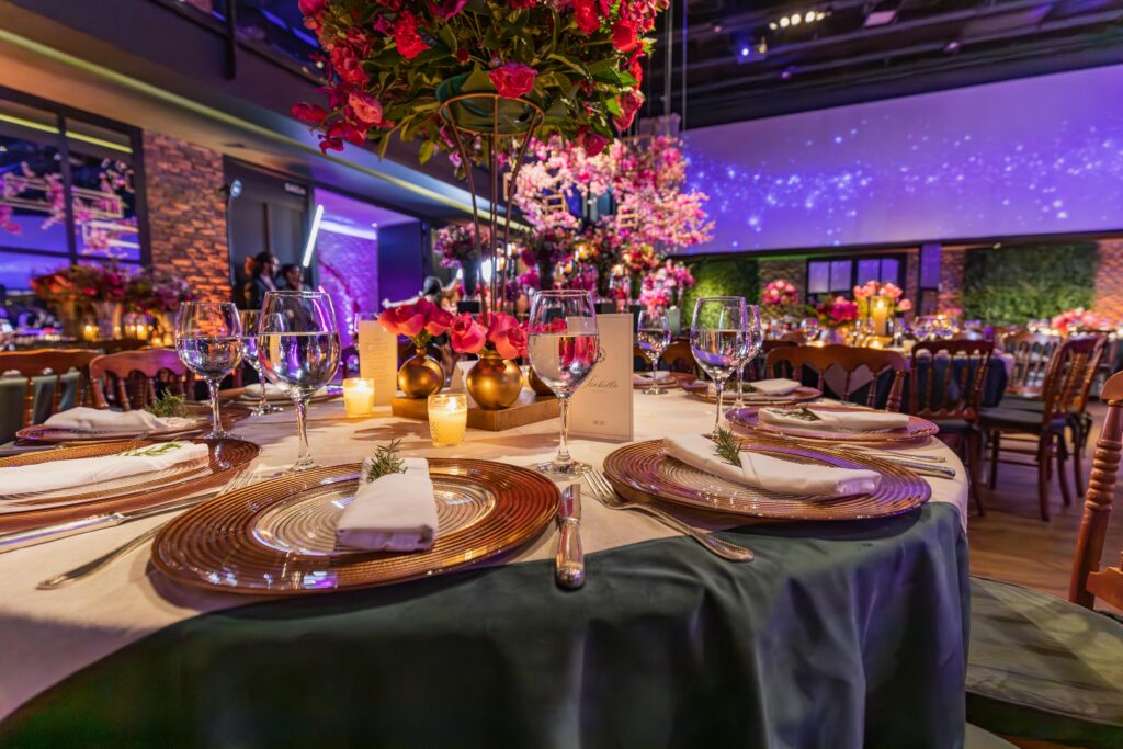 Mesa de boda elegantemente decorada con platos dorados, copas de cristal, servilletas blancas, velas encendidas y un centro de mesa alto con flores rosas, en un salón iluminado con luces tenues y ambiente romántico.