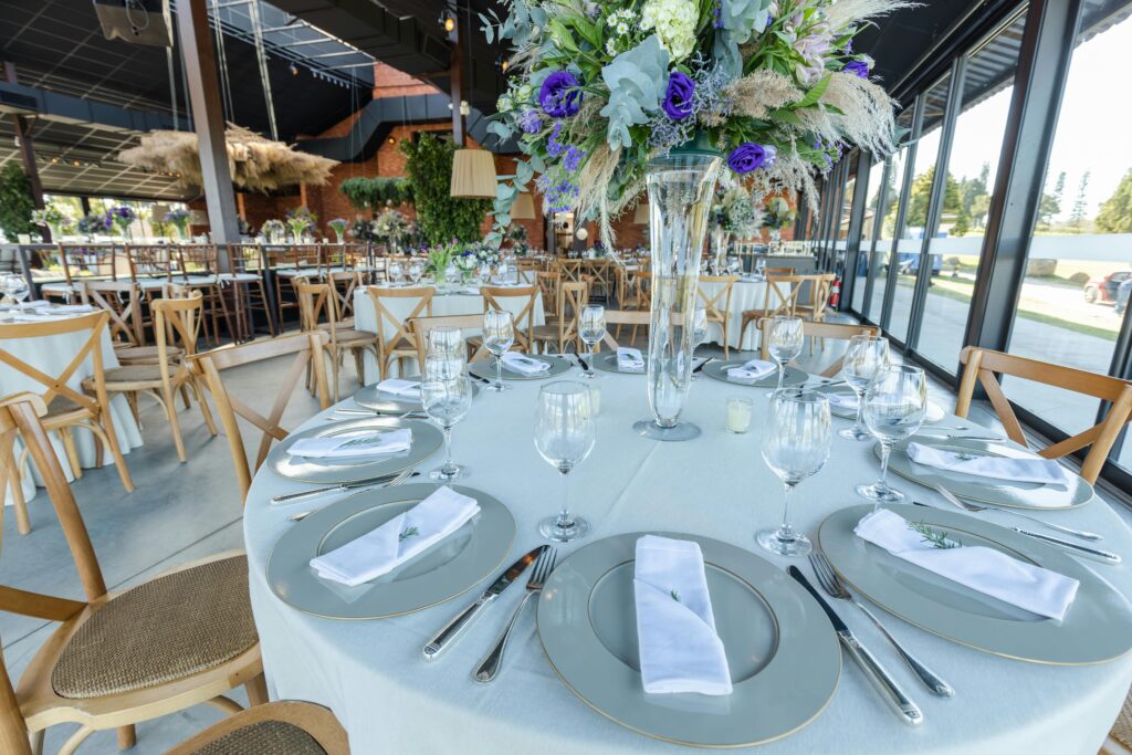 Mesa elegante decorada para recepción de boda, con vajilla en tonos neutros, copas de cristal y centro de mesa floral alto, como parte de un servicio de catering para bodas en Bogotá en un espacio moderno y luminoso.