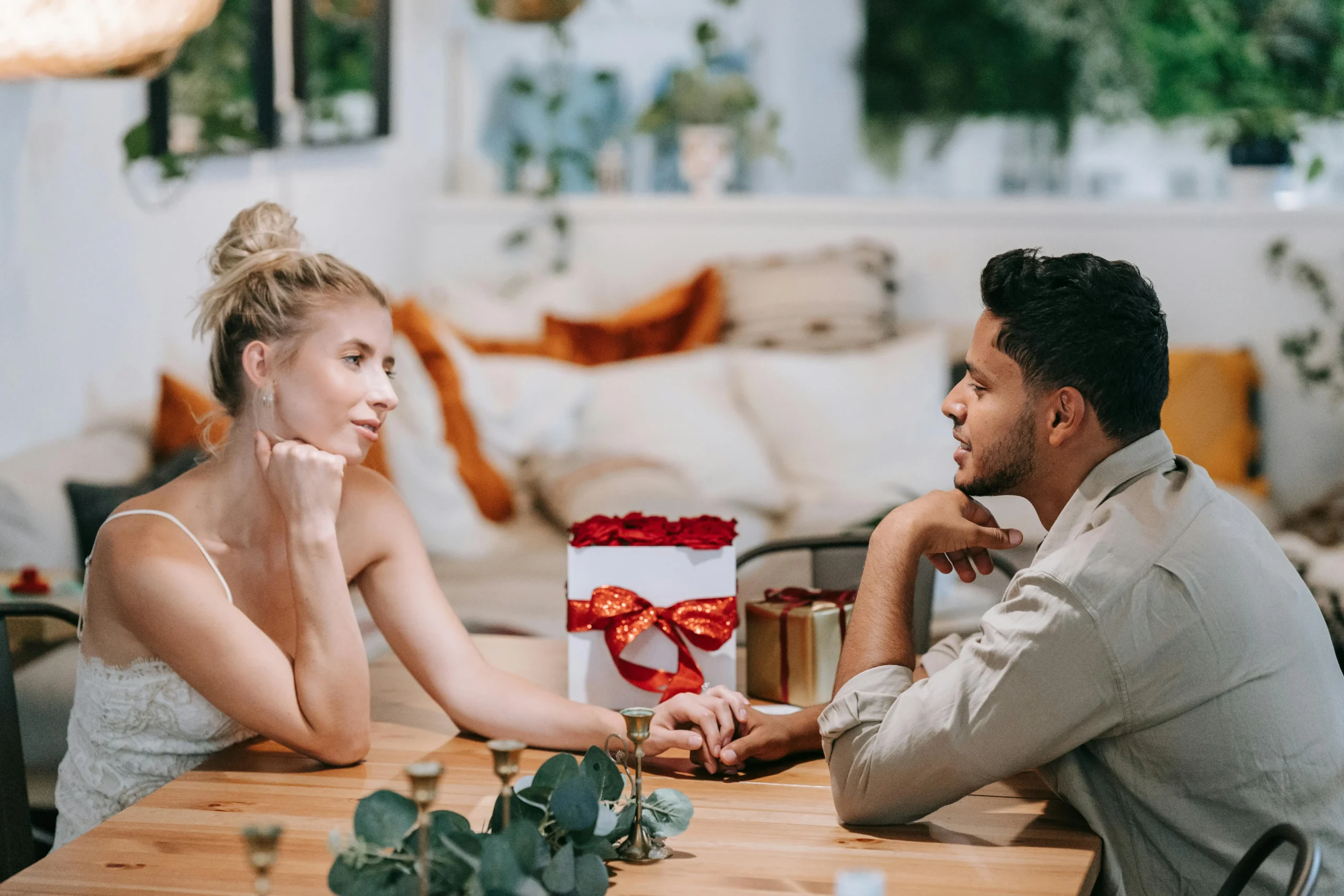 Pareja joven sentada frente a frente en una mesa, tomados de la mano con una caja de regalo entre ellos, en un ambiente romántico de celebración de aniversario