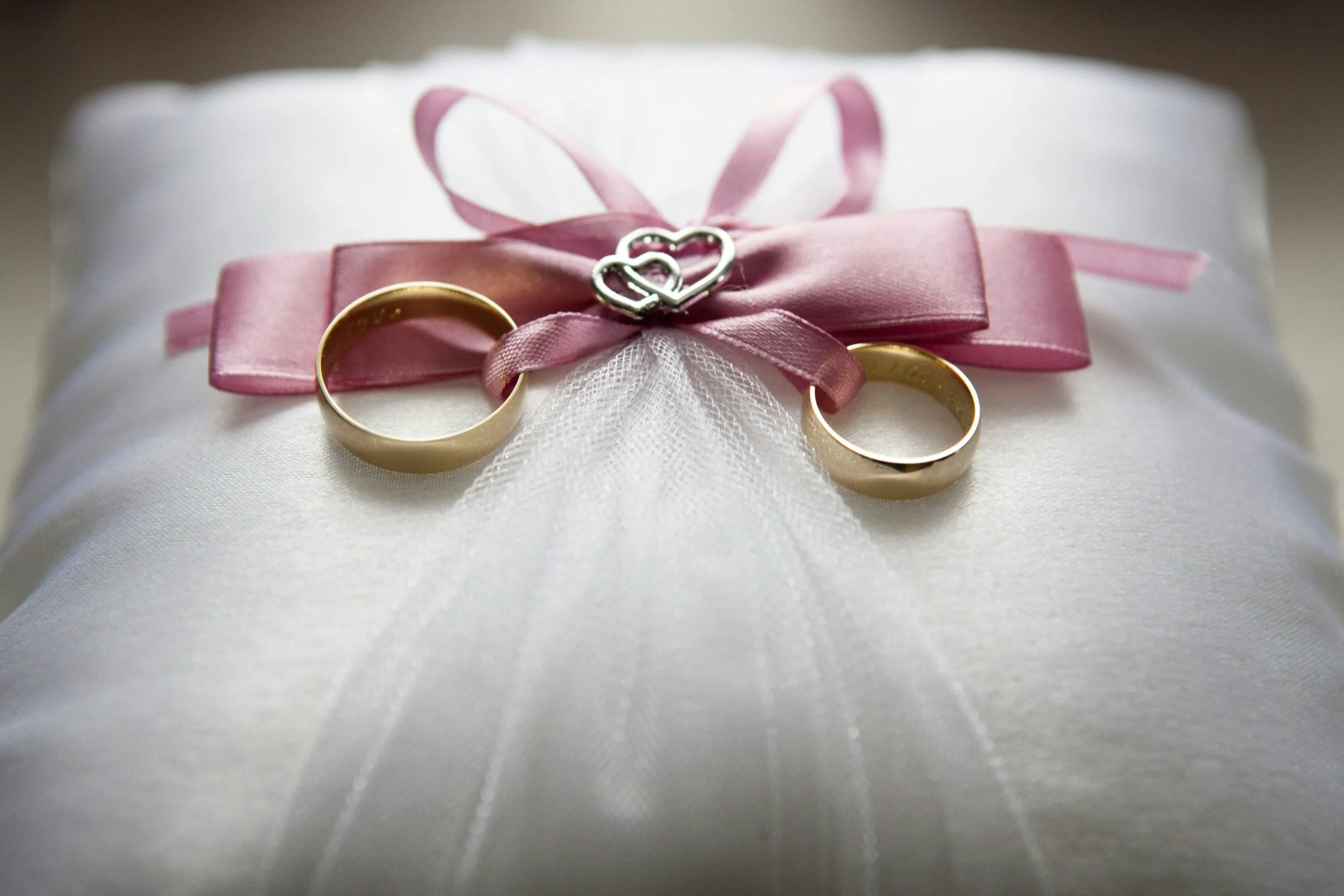 dos anillos de boda dorados sobre un velo blanco, adornados con una cinta rosa y un dije de corazón, simbolizando un aniversario de lino