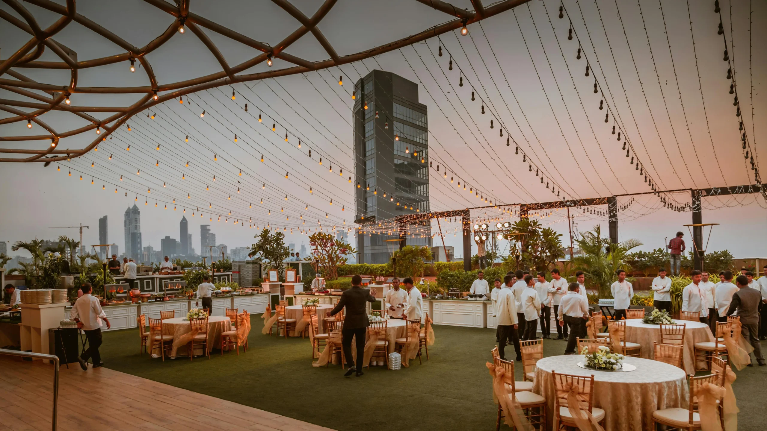 Elegante montaje de evento en terraza de Bogotá con mesas redondas, personal de servicio y guirnaldas de luces, con edificios de la ciudad al fondo