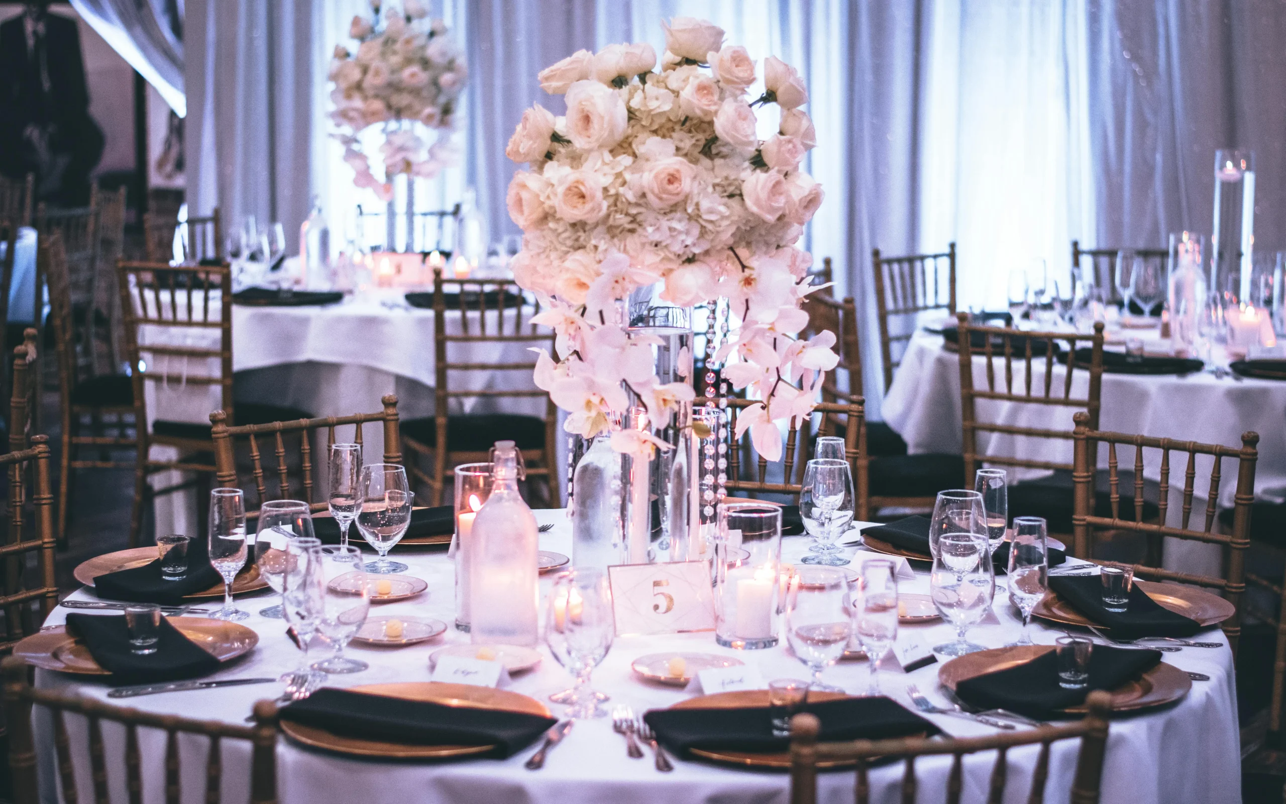 Elegante mesa redonda para celebración con mantel blanco, sillas doradas, y un alto centro de mesa con flores blancas