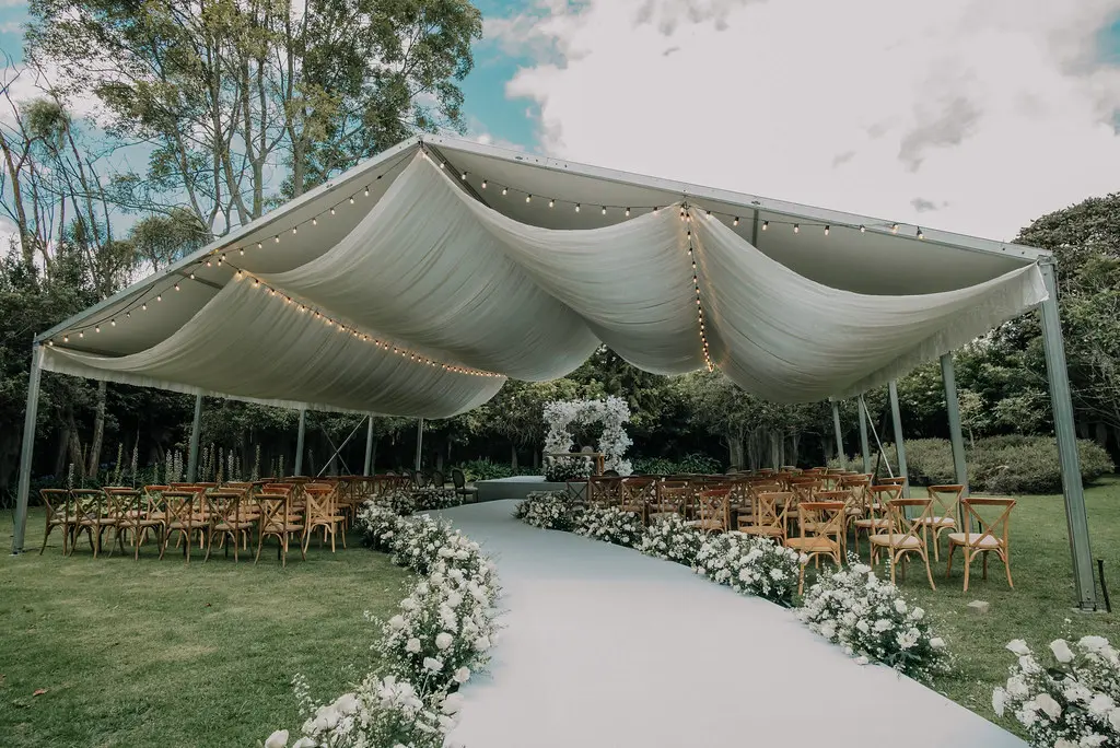 Pasillo de ceremonia de boda al aire libre, decorado con un camino blanco, flores blancas y cubierto por una elegante carpa