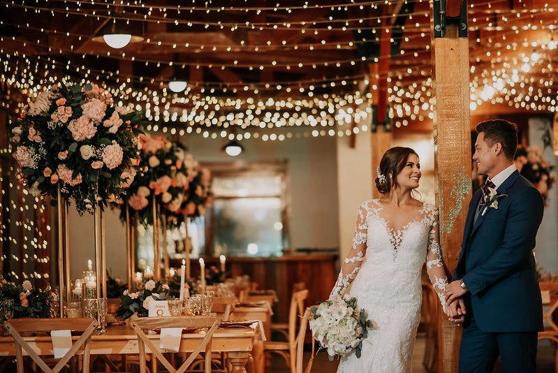 Novios en la recepción de su boda rústica en Subachoque, con decoración de luces cálidas y arreglos florales altos.