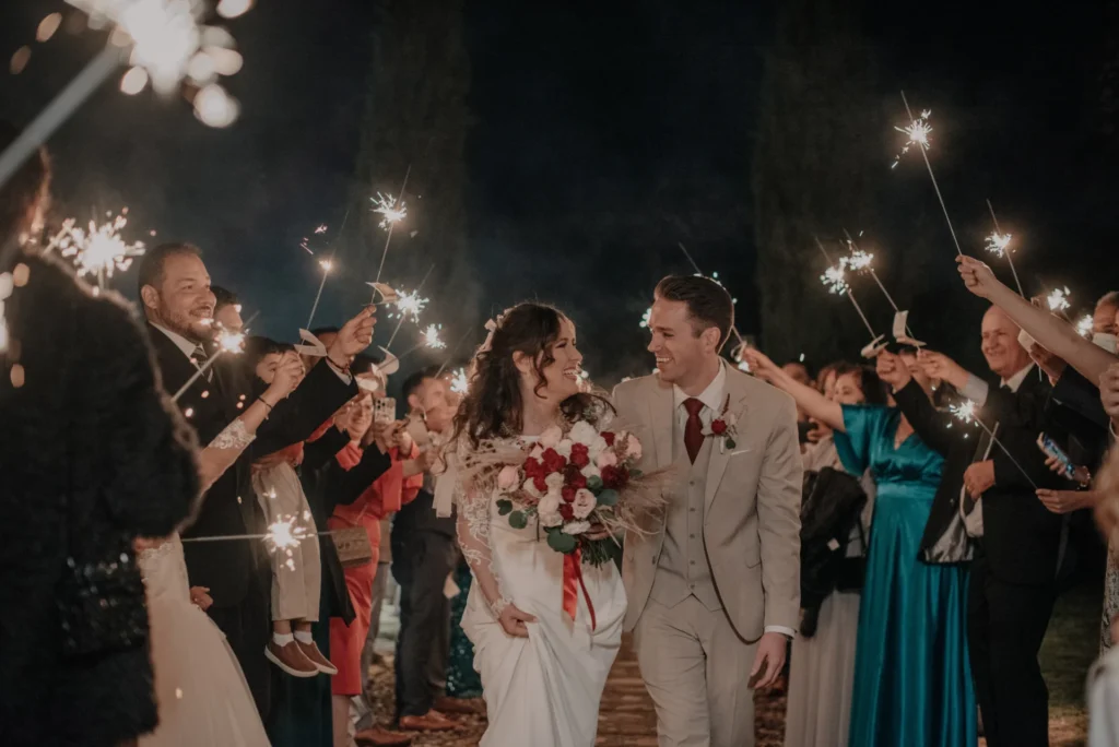 Feliz pareja de novios camina a través de un pasillo de invitados que sostienen bengalas encendidas, celebrando su boda en Zipaquirá. La novia lleva un ramo de rosas rojas y blancas.