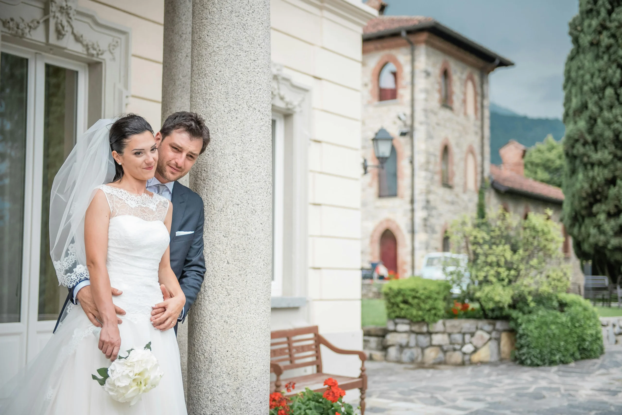 areja de novios posando junto a una columna de piedra durante su boda en Zipaquirá. El novio abraza a la novia, que viste un elegante vestido blanco y sostiene un ramo de flores blancas.