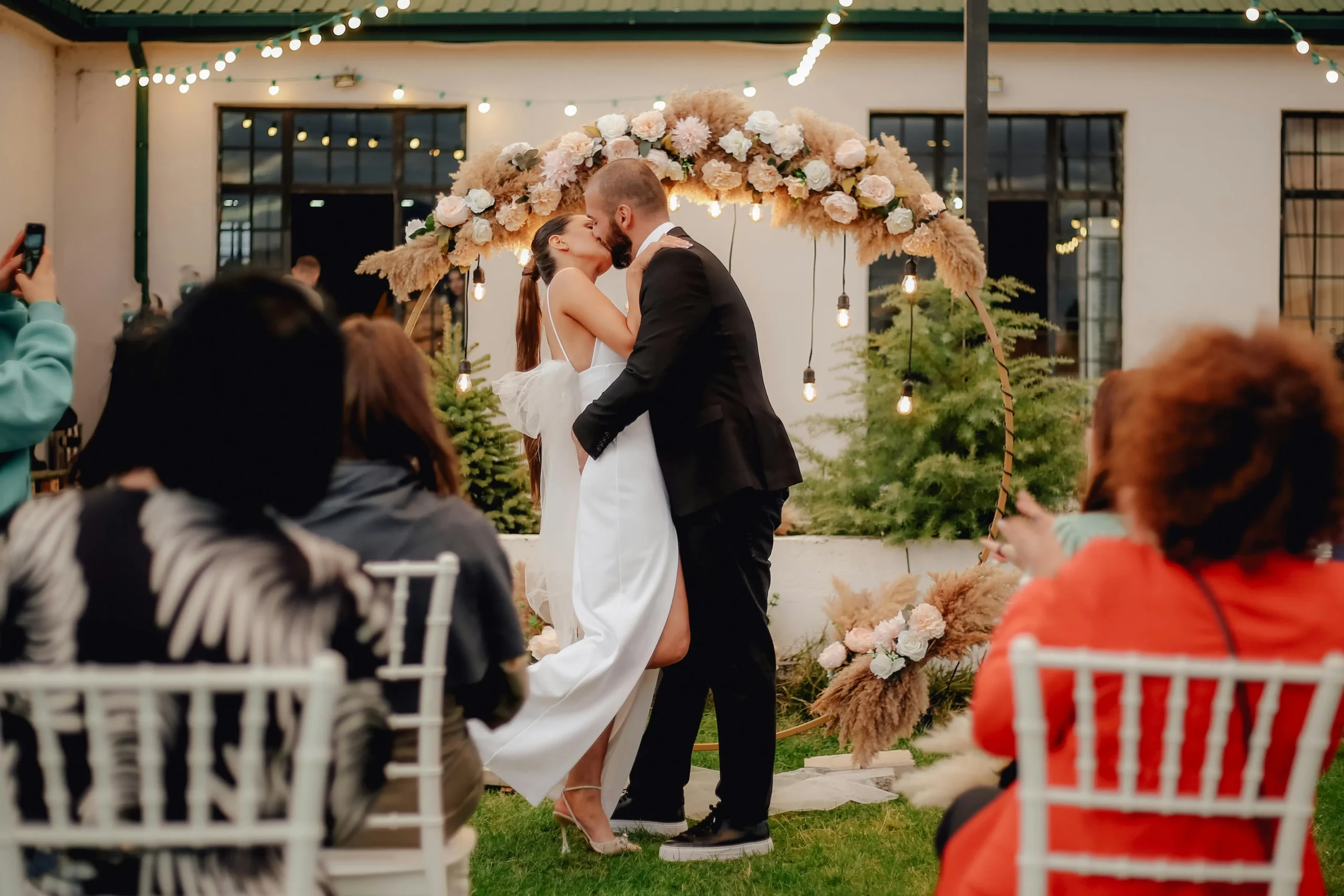 Novios besándose bajo un arco de estilo boho-chic durante su recepción de bodas en Guatavita, rodeados por luces cálidas.