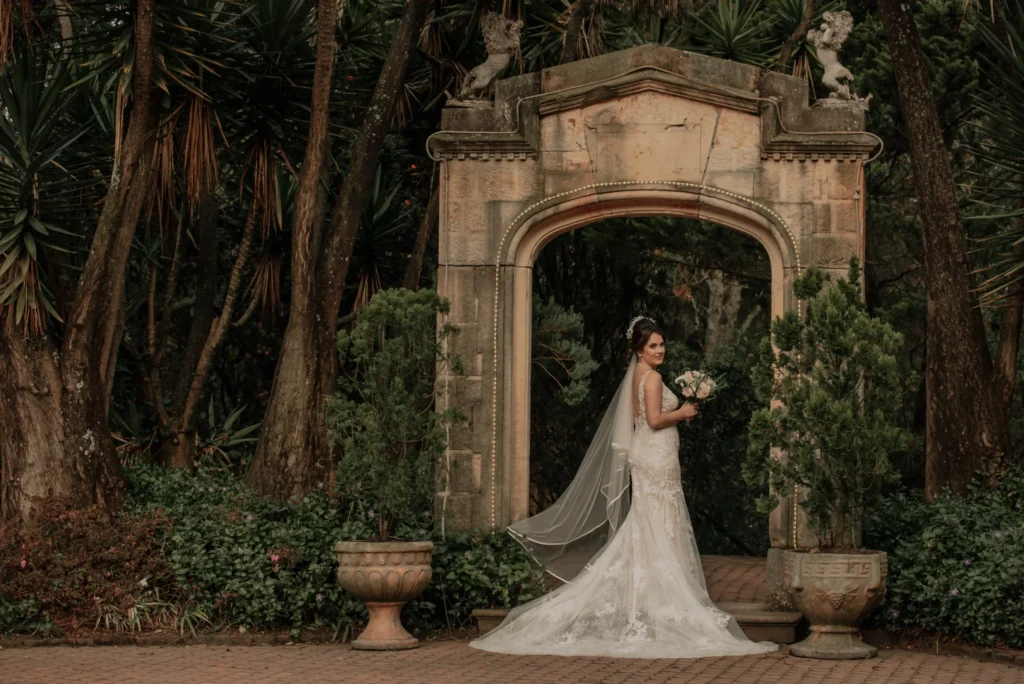 Novia elegante posando bajo un arco de piedra en el jardín de su boda campestre, planificada por Eventos Bella Suiza