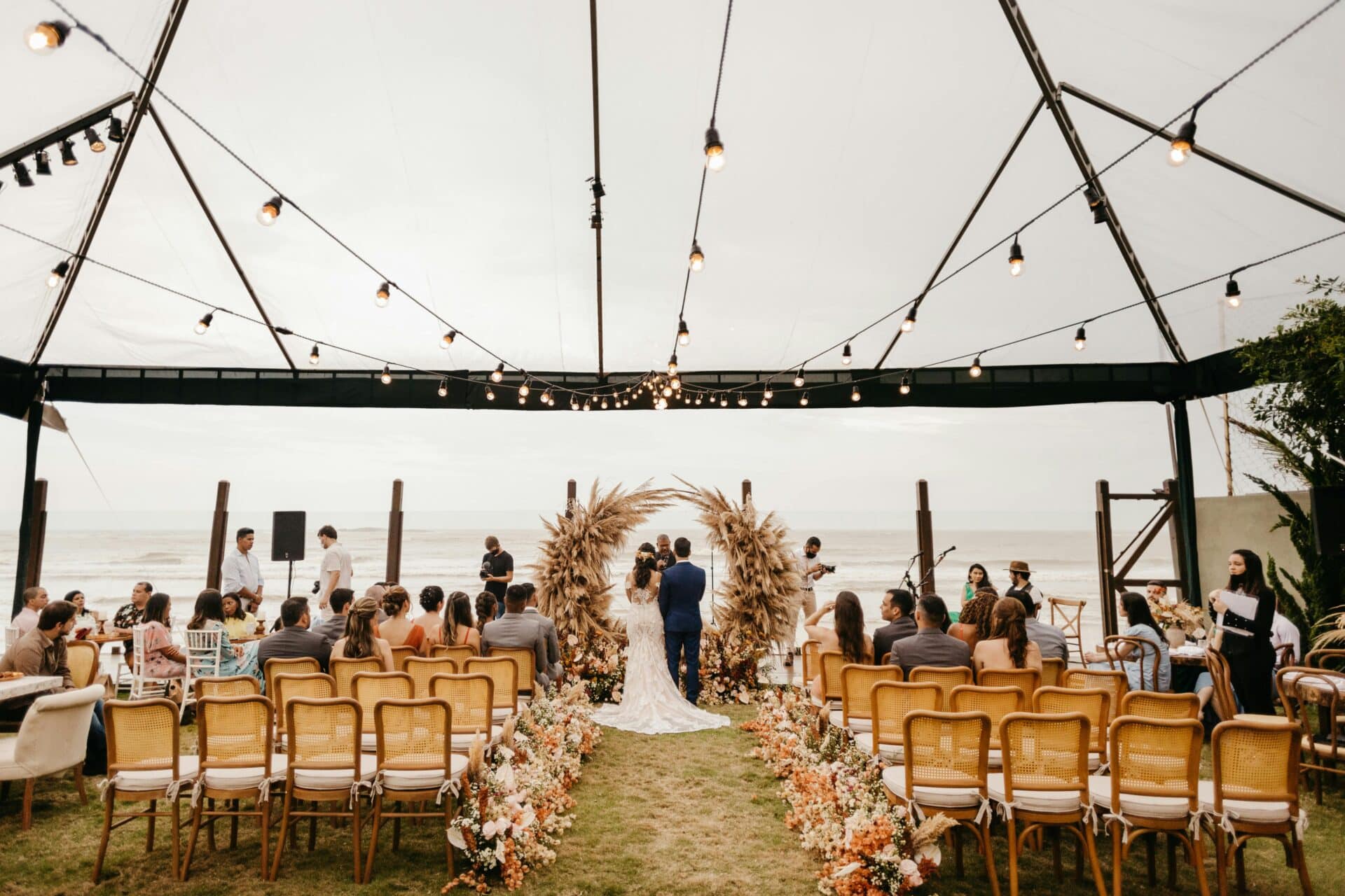 Pareja de novios en ceremonia de boda al aire libre bajo una carpa, con sillas de madera, iluminación de guirnaldas y un arco floral rústico, rodeados de invitados